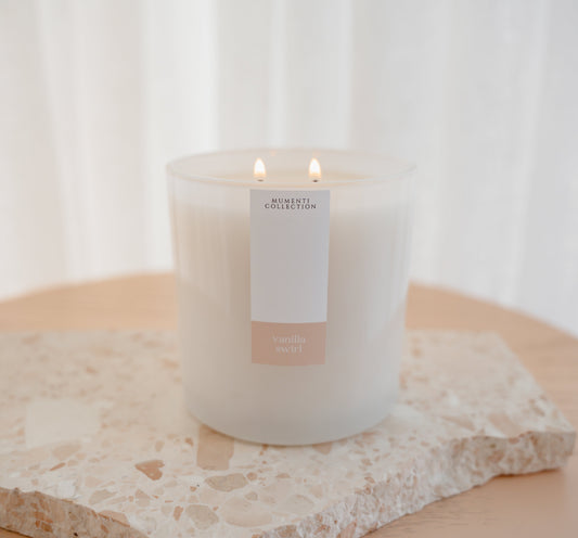 A "Vanilla Swirl" sensory-friendly candle with glowing cotton wood wicks on a quartz stone stand, featuring a white and bright minimalist aesthetic for a home sanctuary.
